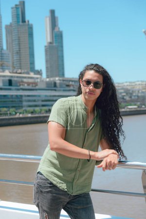Ruhiger junger Latino mit langen lockigen Haaren, Sonnenbrille und gutaussehend, im Urlaub stehend und an Geländer der argentinischen Frauenbrücke gelehnt, Gebäude und Himmel im Hintergrund, vertikales Bild.
