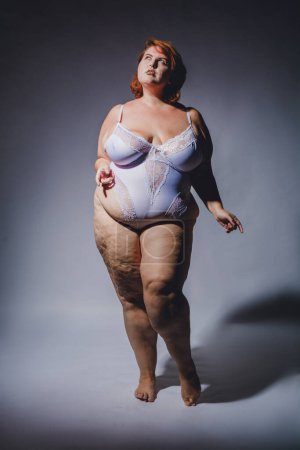 Vertikales Bild der jungen Plus Size Latina Frau argentinischer Herkunft posiert in Dessous mit Cellulite posiert mit Zuversicht, Studiofoto mit hohem Kontrast und hartem Licht.