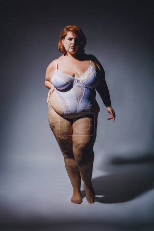 Vertikales Bild der jungen Plus Size Latina Frau argentinischer Herkunft posiert in Dessous mit Cellulite posiert mit Zuversicht, Studiofoto mit hohem Kontrast und hartem Licht.