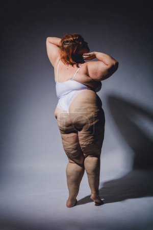 Vertical image of young plus size Latina woman of Argentinian ethnicity posing in lingerie with cellulite posing with confidence, studio photo with high contrast and hard light.