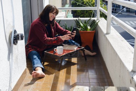 Eine erwachsene Frau argentinischer Abstammung sitzt an einem sonnigen Morgen auf der Terrasse ihrer Wohnung und liest Nachrichten auf ihrem Handy, während sie Bücher liest und Kaffee trinkt.