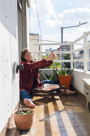 Lateinamerikanische erwachsene Frau auf der Terrasse ihrer Wohnung, die ein Selfie-Foto mit ihrem Handy macht, während sie im roten Pullover sitzt, ruhig Buch liest und Kaffee trinkt, Lifestylekonzept