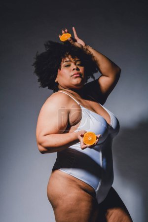 Waist up portrait of young latin afro brunette woman posing in lingerie with orange fruit in hands and looking at camera, studio shot, high contrast light.