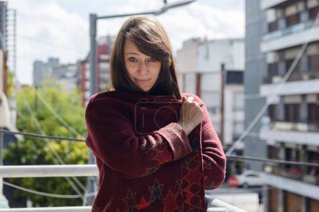 Portrait of adult Latin woman standing in morning on balcony of her bedroom, she covers herself with sweater, she is looking at camera quiet and happy, calm and smiling calmly with city in background.