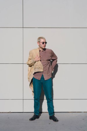 vertical image of young man who is ready to leave, he is standing putting on his jacket, he is wearing sunglasses.