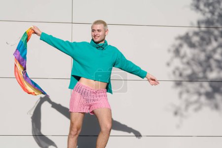 young blond latino man with short hair wearing green sweater and pink shorts dancing with pride flag outdoors, he is very proud for being gay.