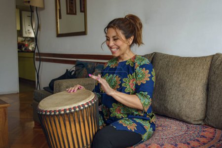 Una mujer latina feliz adulta sentada en casa expresándose a través de la música africana con un tambor Djembe tocando música de percusión africana. Ella sonríe y se divierte.