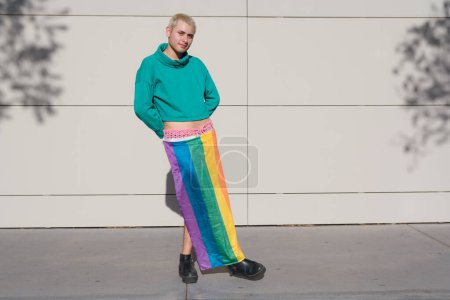 Un homme avec un sweat à capuche vert porte un drapeau de fierté comme une jupe. Il se tient debout devant un mur à midi, regarde la caméra sourire et poser, copier l'espace.