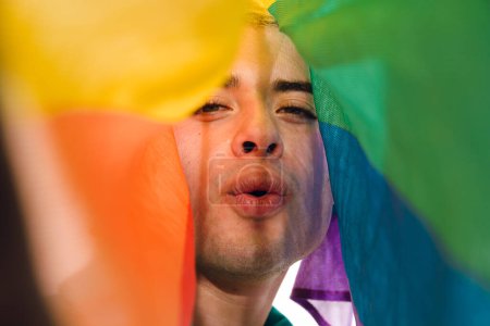 Vista frontal de cerca del Hombre Gay Latino mirando a la cámara soplando un beso cubierto por una bandera de orgullo. Concepto de humor y ligereza, ya que el hombre está haciendo un gesto tonto mientras sostiene la bandera colorida