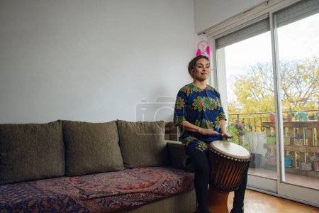 An Adult Woman sitting at home with the eyes closed inspired playing the Djembe drum. he is sitting in a coach by the balcony and the atmosphere seems to be relaxed and casual