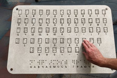 Téléchargez les photos : La main d'un homme lit tactile le braille à l'extérieur sur une tablette avec l'alphabet ukrainien (cyrillique) dans un parc public. Journée mondiale de la vue. Journée mondiale du braille. Concept de la journée des aveugles et des handicapés - en image libre de droit