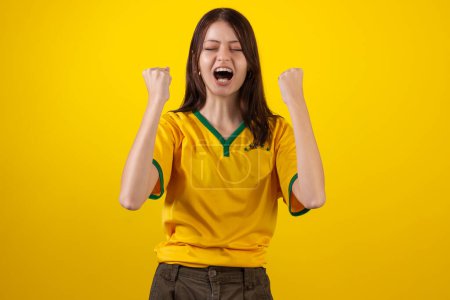 Photo for Oung woman wearing the official uniform shirt of the Brazilian soccer team in the 2022 Qatar Cup and with the flag of Brazil in studio photo. Brazilian fan. - Royalty Free Image