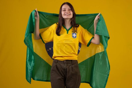 Photo for Oung woman wearing the official uniform shirt of the Brazilian soccer team in the 2022 Qatar Cup and with the flag of Brazil in studio photo. Brazilian fan. - Royalty Free Image