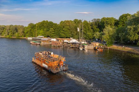 Foto de Berlin-Treptow, Alemania - 9 de junio de 2023, Vista del Spree con restaurantes y varios alquileres de barcos en Treptower Park. Los barcos de excursión van en el río, espacio de copia. - Imagen libre de derechos