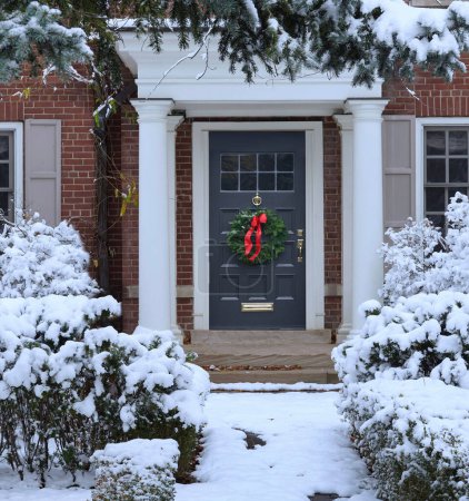 Snow covered pine tree and shrubbery in front of traditional brick house
