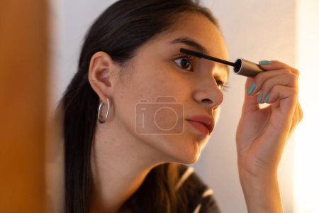 Junge ethnische Dame schaut in den Spiegel, während sie Wimperntusche aufträgt 
