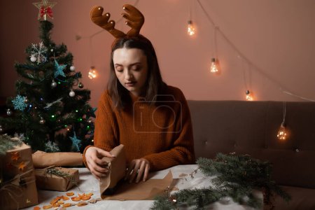 Téléchargez les photos : Une jeune femme heureuse se prépare pour Noël et emballer des cadeaux, la fille a des bois de cerf du Nouvel An sur la tête, un arbre de Noël et des lumières sont en arrière-plan, le concept de préparation pour la nouvelle année, joyeux Noël, nouvelle année 2023 - en image libre de droit