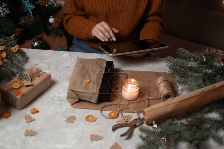 Téléchargez les photos : Une jeune femme tient un téléphone, une tablette à la main et remplit une commande par téléphone, prenant une commande pour emballage cadeau, en arrière-plan un arbre de Noël et des lumières, le concept de préparation pour la nouvelle année, joyeux Noël, nouvel an 2023 - en image libre de droit