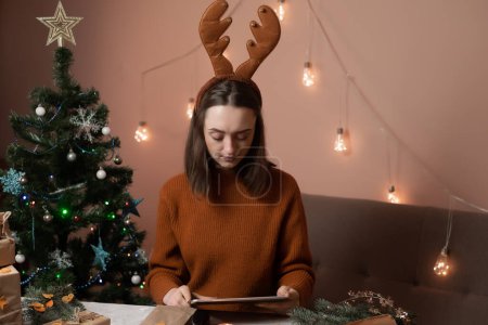 Téléchargez les photos : Une jeune femme tient un téléphone, une tablette à la main et remplit une commande par téléphone, prenant une commande pour emballage cadeau, en arrière-plan un arbre de Noël et des lumières, le concept de préparation pour la nouvelle année, joyeux Noël, nouvel an 2023 - en image libre de droit
