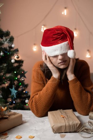 Téléchargez les photos : Fatigué jeune femme dans un chapeau de Nouvel An tiré sur ses yeux en prenant une commande pour emballage cadeau, en arrière-plan un arbre de Noël et des lumières, le concept de préparation pour la nouvelle année, joyeux Noël, nouvel an 2023 - en image libre de droit