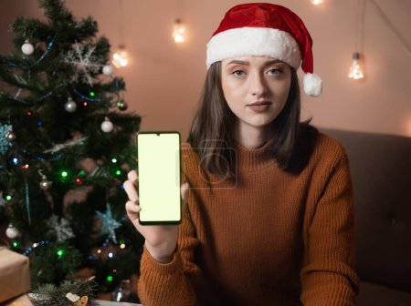 Téléchargez les photos : Une jeune femme heureuse fait une commande par téléphone, la fille a des bois de cerf du Nouvel An sur la tête, prenant une commande pour emballage cadeau, en arrière-plan un arbre de Noël et des lumières, le concept de préparation pour la nouvelle année, joyeux Noël, nouvelle année 2023 - en image libre de droit