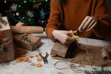 Téléchargez les photos : Gros plan sur les mains d'une fille qui emballe un cadeau de Noël ou d'anniversaire sur une table en béton gris, en arrière-plan un arbre de Noël et des lumières, le concept de préparation pour la nouvelle année, joyeux Noël, Nouvel An 2023 - en image libre de droit
