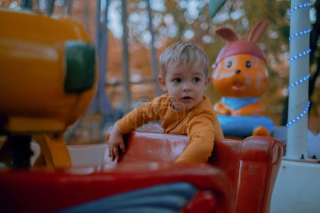 In the autumn park, a little blond boy clad in a yellow sweater joyfully rides the children's carousel. His bright laughter rings out amidst the colorful autumn foliage