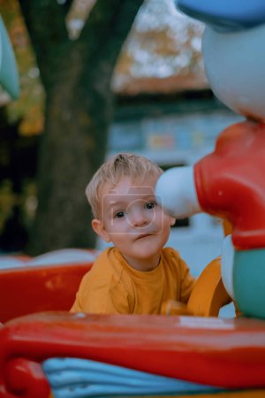 In the autumn park, a little blond boy clad in a yellow sweater joyfully rides the children's carousel. His bright laughter rings out amidst the colorful autumn foliage