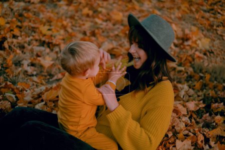 Eine Mutter und ihr Sohn verbringen viel Zeit miteinander und spielen im herbstlichen Park, beide in gelben Gewändern. Inmitten des bunten Laubes teilen sie freudige Momente, Lachen hallt in der klaren Luft wider.