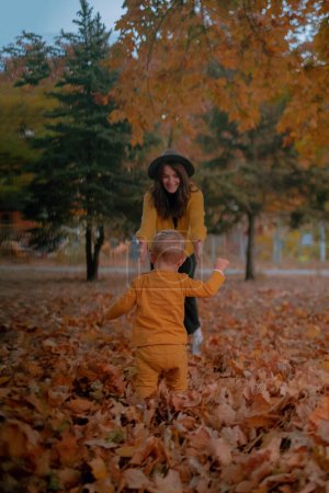 Eine Mutter und ihr Sohn verbringen viel Zeit miteinander und spielen im herbstlichen Park, beide in gelben Gewändern. Inmitten des bunten Laubes teilen sie freudige Momente, Lachen hallt in der klaren Luft wider.