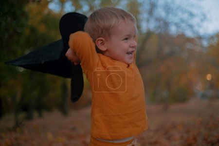 Un niño pequeño se sienta en un sombrero negro de Halloween, sosteniendo una calabaza, sonriendo en medio de hojas de otoño en el parque.
