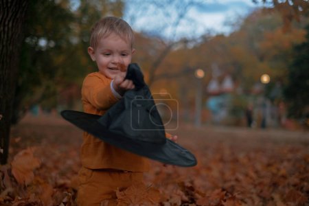 A little boy sits in a black Halloween hat, holding a pumpkin, smiling amidst autumn leaves in the park.