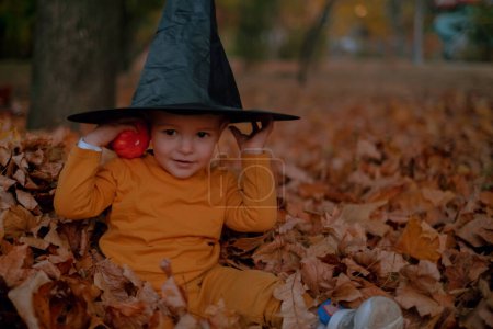 Ein kleiner Junge sitzt in einem schwarzen Halloween-Hut, hält einen Kürbis in der Hand und lächelt inmitten von Herbstblättern im Park.