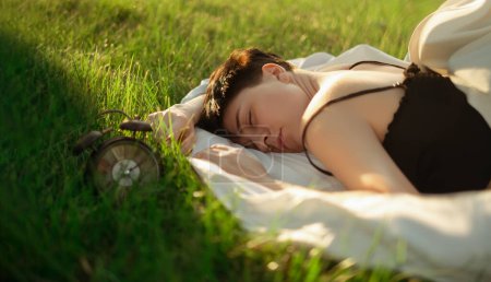 A girl wrapped in a blanket lies on green grass next to an alarm clock, promoting healthy sleep habits and the importance of waking up refreshed with the help of an alarm clock