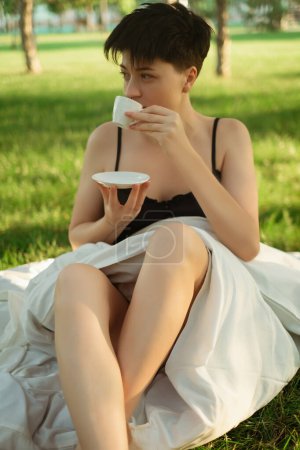 Ein in eine Decke gehülltes Mädchen sitzt mit einer Tasse Kaffee auf grünem Gras und genießt ein erfrischendes Morgengebräu. Coffeeshops im Park bieten einen herrlichen Start in den Tag mit belebendem Morgenkaffee.