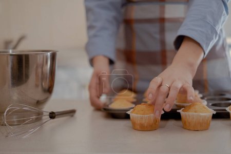 Une boulangère tient des cupcakes fraîchement cuits, mettant en valeur sa pâtisserie maison. Embrassez l'art de la cuisson à la maison avec cette image d'un chef pâtissier qualifié travaillant à la maison.