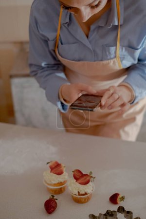 A female pastry chef takes photos on her mobile phone to advertise her business, highlighting homemade baking. Experience the essence of small-scale, eco-friendly baking with gluten-free and sugar-free products, promoting healthy eating.