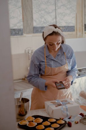 A female chef- pastry chef takes photos on her mobile phone to advertise her business, highlighting homemade baking. Experience the essence of small-scale, eco-friendly baking with gluten-free and sugar-free products, promoting healthy eating.