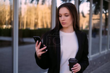 A businesswoman in a suit holding a phone and coffee, office worker, social media manager, freelancer, outdoor workspace, remote work, modern professional, multitasking.