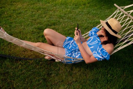 Girl sits in hammock, working on phone while on vacation. Remote work and freelancing, technology and mobile apps, travel and work accessories, time management, and task organization.