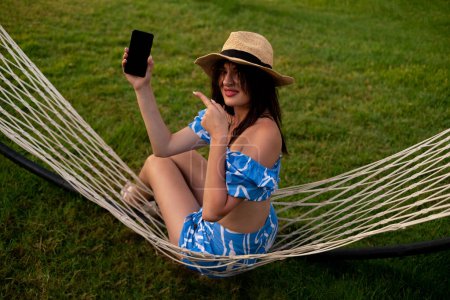Girl points at phone with blank screen, working outdoors from hammock. Remote work, freelancing, technology, mobile apps, travel, work accessories, time management, and task organization.