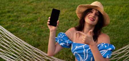 Girl points at phone with blank screen, working outdoors from hammock. Remote work, freelancing, technology, mobile apps, travel, work accessories, time management, and task organization.