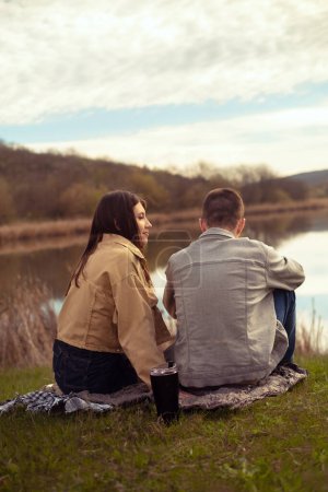 Photo for A young couple picnics by the lake, sipping hot tea and embracing, using a thermos for camping and pouring hot coffee. Valentine's Day - Royalty Free Image