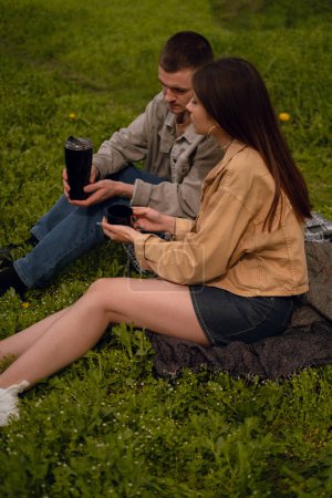 A young couple on a picnic sips hot tea from a thermos, enjoying the convenience and practicality for an active outdoor experience. Refreshments in the open air create a cozy picnic atmosphere.