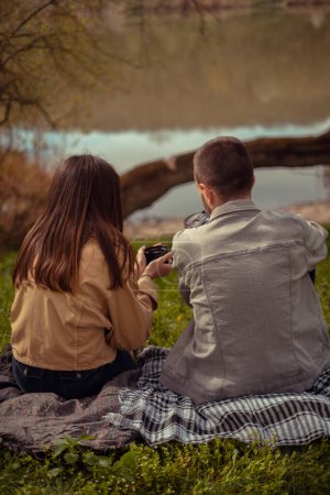 Eine Picknick-Szene am See, während junge Camper heißen Tee genießen. Der Fokus liegt auf dem Rücken eines jungen Paares, das in die Natur eintaucht und den Moment genießt. Valentinstag
