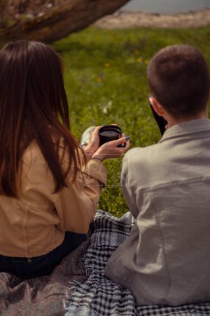 A young couple sits with their backs to the camera by the lake shore, enjoying a picnic in the park and sipping hot drinks. The girl holds a cup of tea, warming her hands.