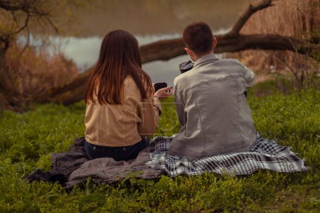 A man and a woman sit by the lake shore with their backs to the camera, enjoying camping in nature in springtime, sipping hot coffee outdoors.