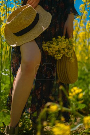Woman's legs in blooming rapeseed field, holding straw hat and purse, immersed in vibrant yellow blossoms, epitomizing summer charm.