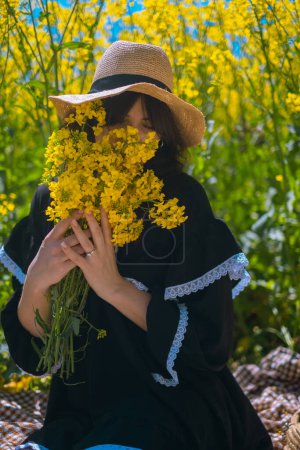 Frau beim Picknick hält Blumenstrauß, trägt Strohhut, im Hintergrund blühendes gelbes Rapsfeld.
