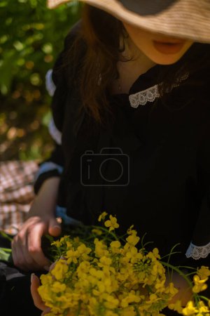 Femme au pique-nique tient bouquet de fleurs, portant un chapeau de paille, avec champ de colza jaune en fleurs en arrière-plan.